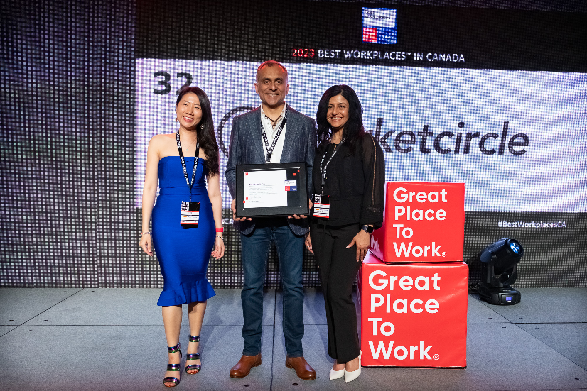 Three people pose in front of a screen during the Best Workplaces in Canada ceremony. They wear formal attire and smile side-by-side. Alykhan Jetha, CEO of Marketcircle, stands in the middle holding Best Workplaces in Canada award.